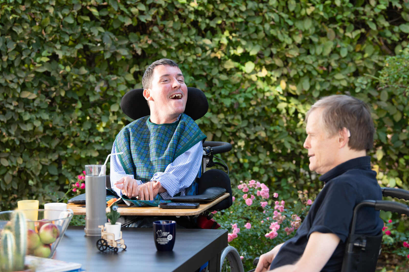 Zwei Männer im Rollstuhl sitzen im Garten an einem Tisch mit Obst und Tassen.
